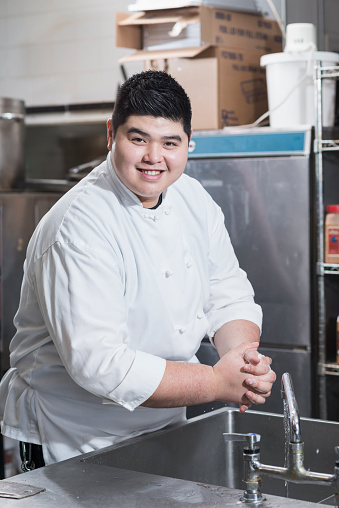 A young Asian man working in a restaurant, washing his hands in the stainless steel sink in a commercial kitchen.  He is wearing a white chef's uniform, smiling at the camera.