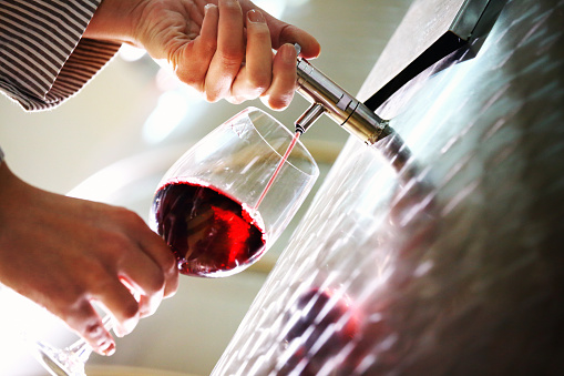 Closeup of unrecognizable adult poouring red wine into glass. Employee of the local winery, filling the glass for tasting. Wine is stored in stainless steel wine tanks