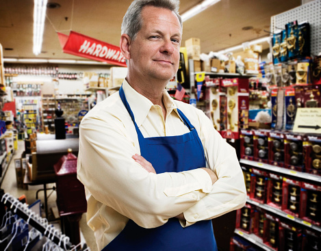 Portrait of middle age hardware store manager wearing blue apron with customers in the background.