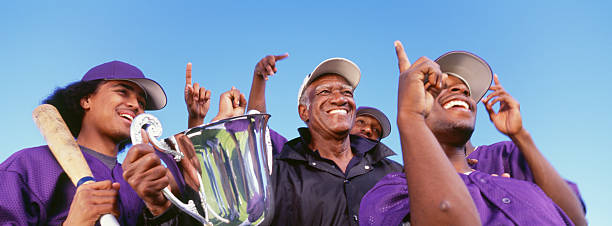 Panoramic shot of cheerful baseball players with trophy Panoramic shot of cheerful baseball players with trophy celebrating victory against clear sky men baseball baseball cap baseball bat stock pictures, royalty-free photos & images