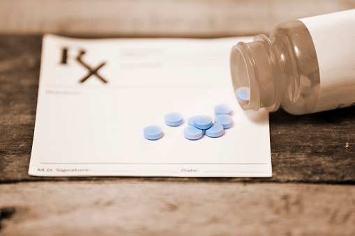 White, blank doctor's prescription pad with a vintage, sepia-toned prescription medication bottle lying on top of it with pills spilling out of the bottle.  The bottle has a blank label on it.  Blue colored pills lay on top of prescription.  A large \