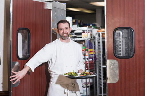 A chef or waiter carrying a tray of food from the kitchen, walking through a doorway.  He is smiling at the camera, wearing a white uniform and apron.