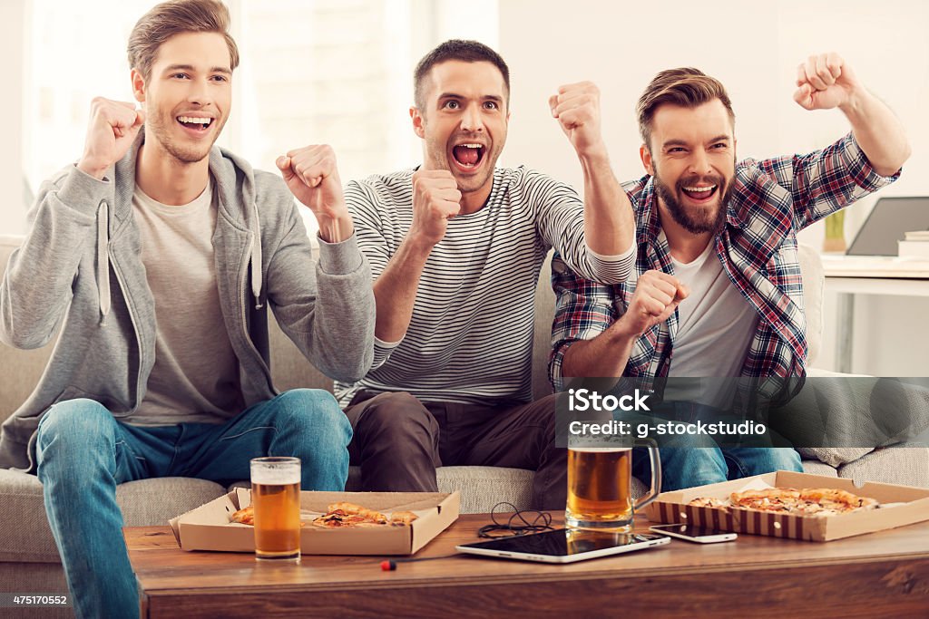 Domestic fans. Three happy young men watching football game and keeping arms raised while sitting on sofa American Football - Sport Stock Photo