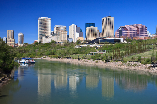 Downtown buildings of Edmonton, Alberta, Canada. The city of Edmonton is the capital city of Alberta. It is a city of nearly one million people. Image is taken on a picture perfect spring day when greens are coming to life. There are no clouds in the sky and the downtown skyscrapers stand in the distance. Image is taken near the North Saskatchewan River in one of many parks. A riverboat, also an attraction in the city, is in the foreground. Buildings are reflected in the river.