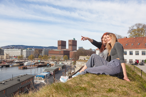 Two beautiful nordic girls enjoying their spare time in Oslo, talking and laughing, with harbour and town hall on background. Lifestyle and friendship concepts.