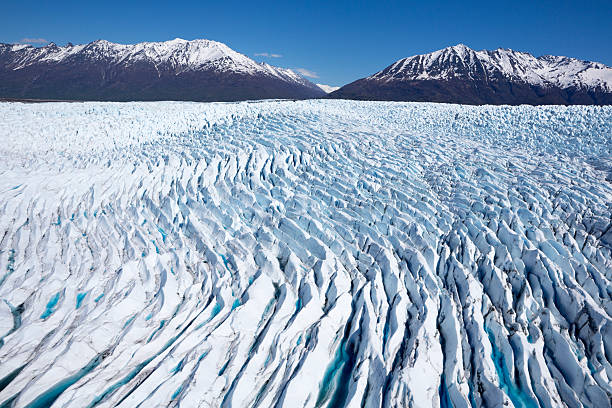 The Knik River and its Knik Glacier or Matanuska Glacier are starting to thaw and break apart.  The ice is a blue color, with the Alaska Mountain Range is the source of the river flow.  Ice, water and dirt are seen in the thawing ice and snow.