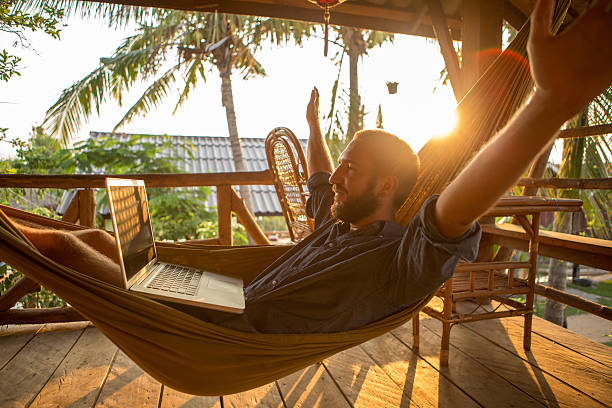 Young man lying down on a hammock at sunset with his computer. Arms outstretched for freedom and positive emotion.