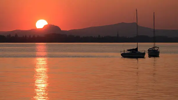 The sun goes under over the Lake of Constance. In the foreground, the Lake of Constance with two sail vessel and in the background the volcano mountains of the "Hegau" with the "Hohentwiel".