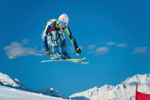 Front view of young male skier at straight downhill race in mid-air against the bue sky and the mountains