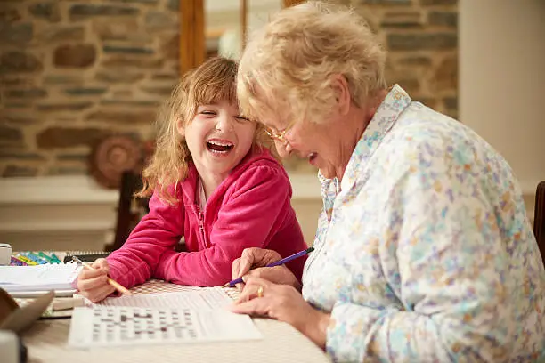 A 5 year old girl is writing in a puzzle book with her grandma and laughing together . They are sitting at a dining table and holding pencils. There is a puzzle crossword book on the table .