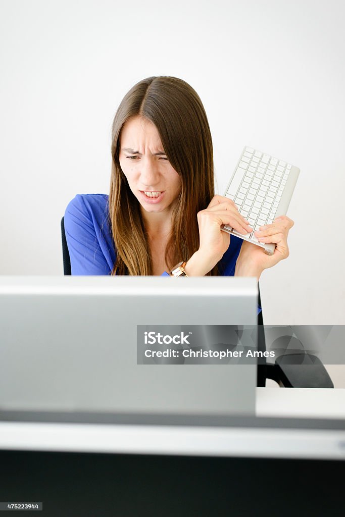 Angry Female Office Worker Attacks Computer Screen With Keyboard An attractive young woman with long brown hair angrily attacks her computer monitor using the computer's keyboard. Camera: 36MP Nikon D800E. 2015 Stock Photo