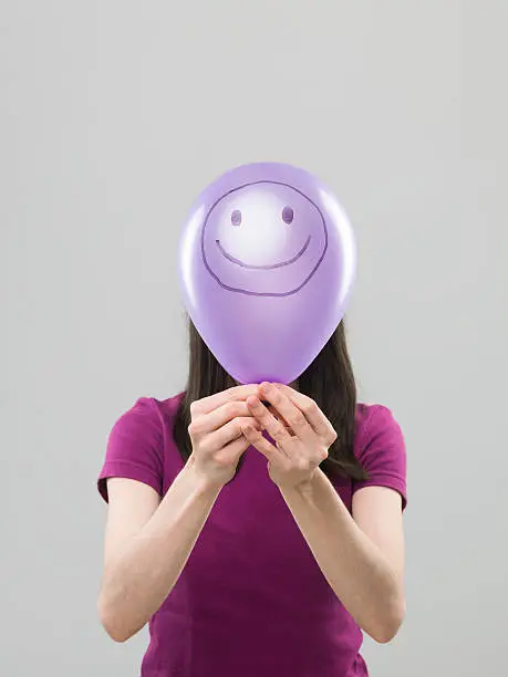 woman hiding her face behing purple balloon with smiley face drawn on it, on grey background