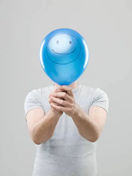 man hiding his face behing blue balloon with smiley face drawn on it, on grey background