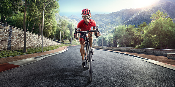 An athlete is riding a bicycle on a spiral track high in the mountains.  The man is wearing black bike shorts and shin guards along with a red sleeveless top and a red and white helmet and sunglasses.