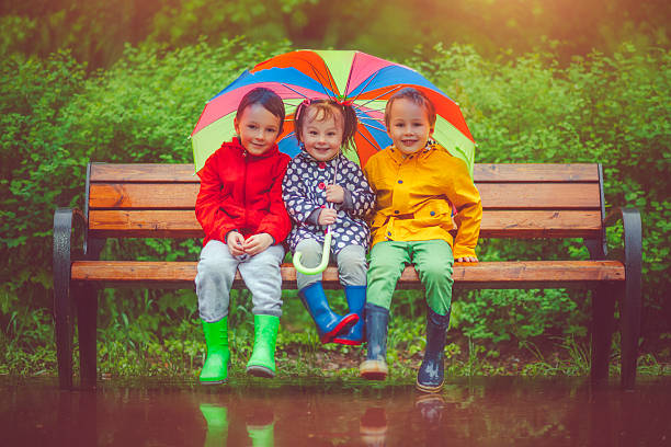 Chilren under umbrella Little boys and girl happily sitting under umbrella in park raincoat photos stock pictures, royalty-free photos & images