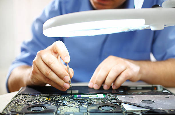 Man unscrewing motherboard of a laptop. Adult caucasian man unscrewing motherboard on a laptop. He's an employee at computer maintenance and service shop. He's using standing magnifying glass with light, The man is wearing blue shirt with rolled back sleeves. installing laptop ram stock pictures, royalty-free photos & images