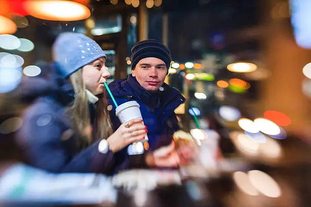 Young tourists, couple, man and woman, drinks coffe in cafe in Manhattan. View trough the window. High ISO, noise, LensBaby photo with selective focus, with reflections.