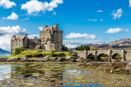 Eilean Donan Castle, Dornie, Scotland - September 20, 2014: colourful view during low tide in a sunny and bright day as wind waves the Scottish flag. Tourists pay visit when touring Highland and Skye.