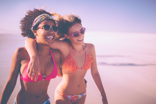 Mixed race girl friends hugging and walking together along a beach on a bright summer day