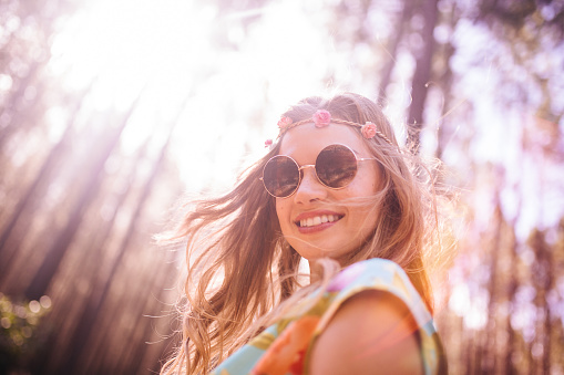 Portrait of a smiling boho girl wearing a flower headband and vintage round sunglasses in a bright forest