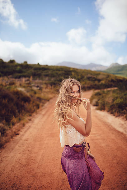 Young smiling girl standing in the middle of an asphalt road holding her longboard against a cloudy sky at sunset