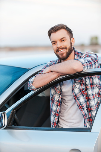 Happy young man leaning at the door of his car and smiling at camera while standing outdoors