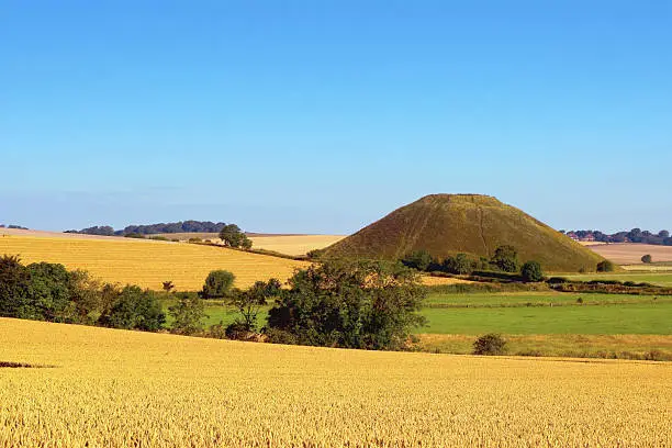 Fields of golden wheat and the distant Silbury Hill with blue sky above. Silbury Hill is part of the Avebury World Heritage Site its purpose and significance remain unknown, the largest man-made mound in Europe.