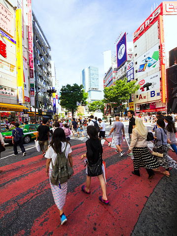 People at motion on crossing not far from Shibuya train station in Tokyo. In background there are surrounding buildings, same trees, commercials and blue, cloudy sky. Shibuya is shopping part of Tokyo and is known for the most crowded crossing in Tokyo, Japan. It is also known for in front of dog (Inu) meeting place.  