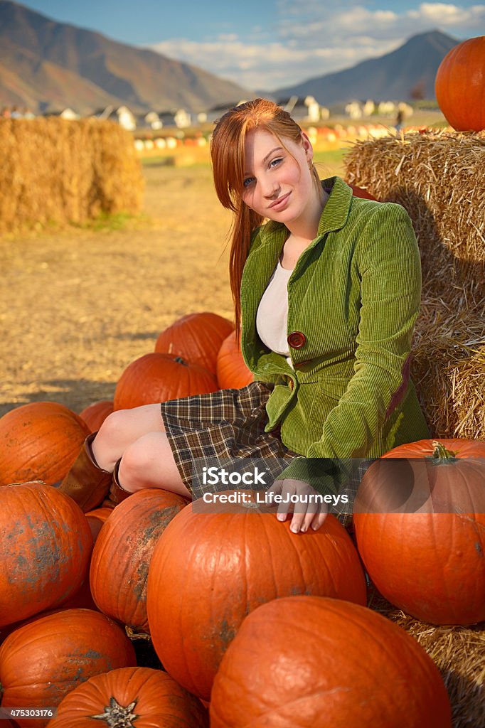 Red Headed Young Woman Sitting On a Pile of Pumpkins Long red haired young woman sitting outdoors on a pile of orange pumpkins.  Smiling and looking at the viewer.  She is wearing a green courduroy jacket, a brown plaid skirt and brown boots. 2015 Stock Photo