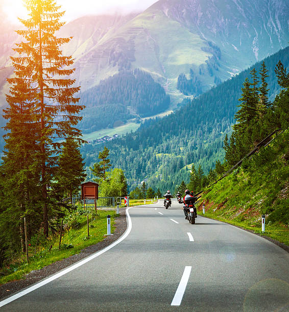 Group of bikers in Alps, active vacation, curve road in the mountains, fresh pine trees along highway, bright sunshine, extreme transportation