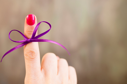 Purple social awareness ribbon tied in a bow around a woman's index finger.  The purple ribbon represents various causes including: domestic violence, various types of cancer, ADD/ADHD, Alzheimer's Disease, animal abuse, Crohn's Disease, Cystic Fibrosis, Fibromyalgia, Epilepsy, and Lupus. Gray background. Copyspace. 