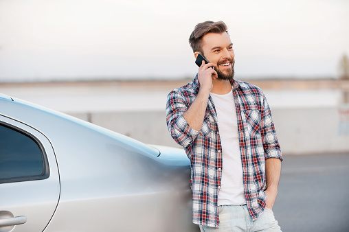 Handsome young man talking on the mobile phone and smiling while leaning at his car