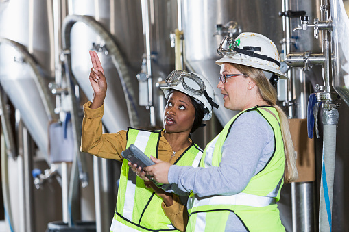 Two multiracial women working together in a manufacturing plant, standing in front of steel storage tanks.  They are wearing white hardhats with safety goggles, and yellow reflective vests.  The African American woman is talking at pointing upward.  The other one is holding a digital tablet.