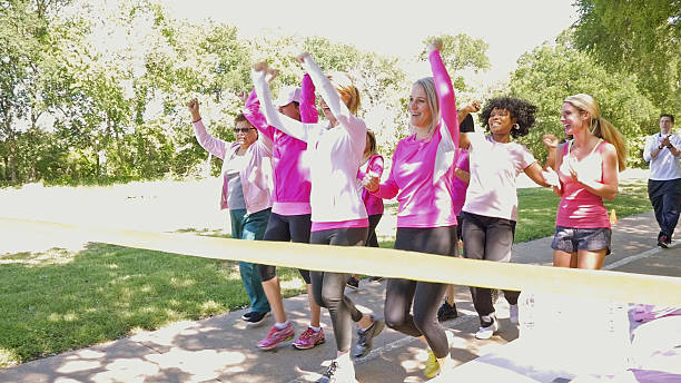 Diverse team of women winning marathon for breast cancer awareness Diverse team of adult women are walking across finish line during marathon or 5k charity race. They are celebrating their success while completing their goal. Runners are wearing pink athletic clothing and breast cancer awareness ribbons. walking for a cause stock pictures, royalty-free photos & images