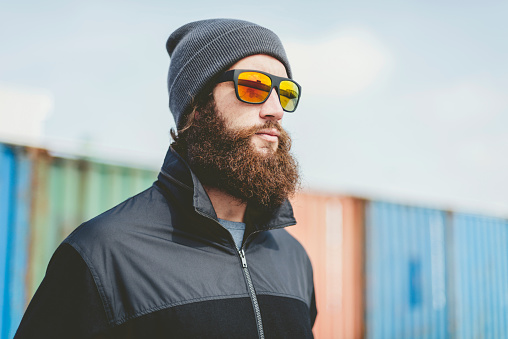 Close up Serious Young Man with Long Beard in Black Jacket and Bonnet with Sunglasses, Looking to Far Right Seriously.