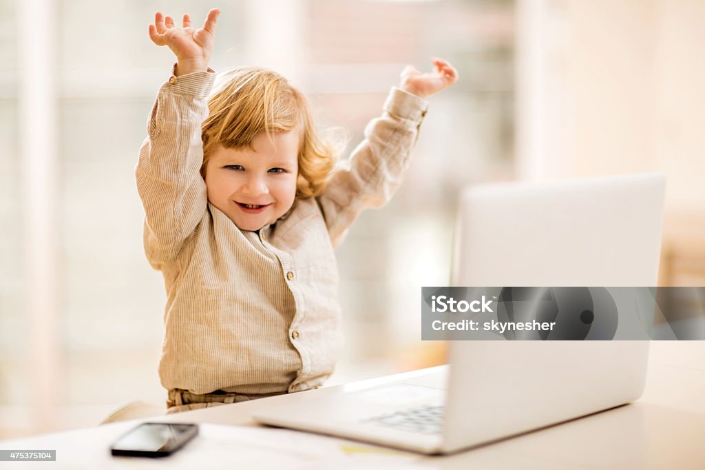 Cheerful business boy celebrating in office with his arms raised. Portrait of cute happy little business boy sitting in the office and celebrating his success with his hands raised. He is looking at camera. Celebration Stock Photo