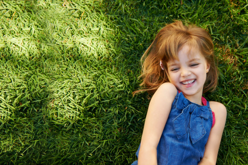 Young blonde caucasian girl in jeans jacket sitting on meadow at sunset time looks at camera happily enjoying outdoor leisure. Child at nature. Happy childhood, eco life.