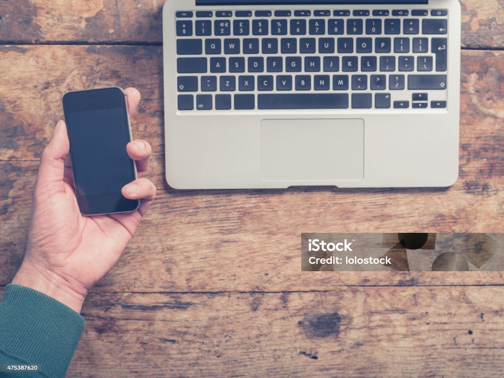 Man using laptop and smartphone Close up on the hands of a young man as he is using a laptop and a smart phone at a wooden table 2015 Stock Photo