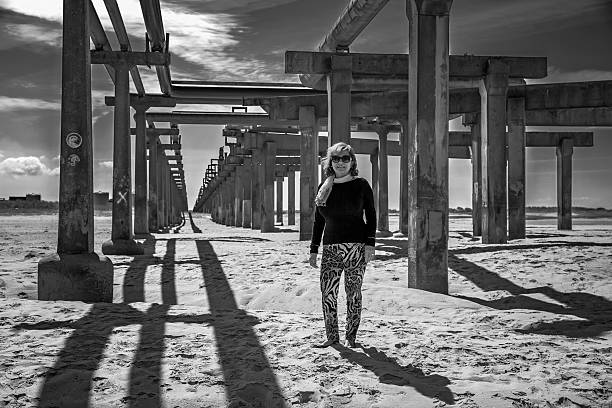 Mature hispanic woman standing on industrial beach amongst pillars holding elevated pipeline