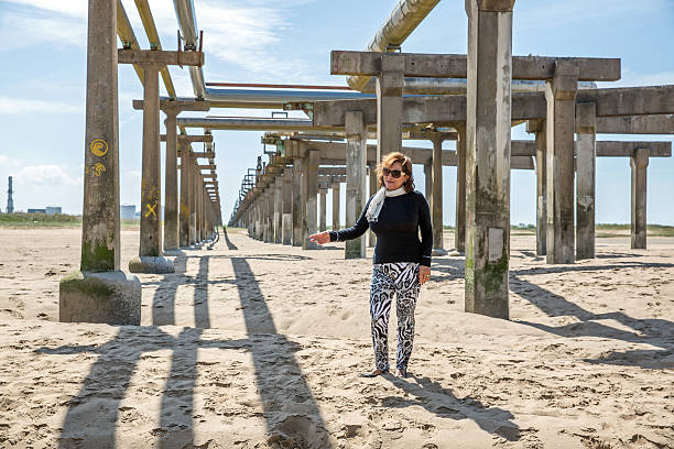 Mature hispanic woman standing on industrial beach amongst pillars holding elevated pipeline