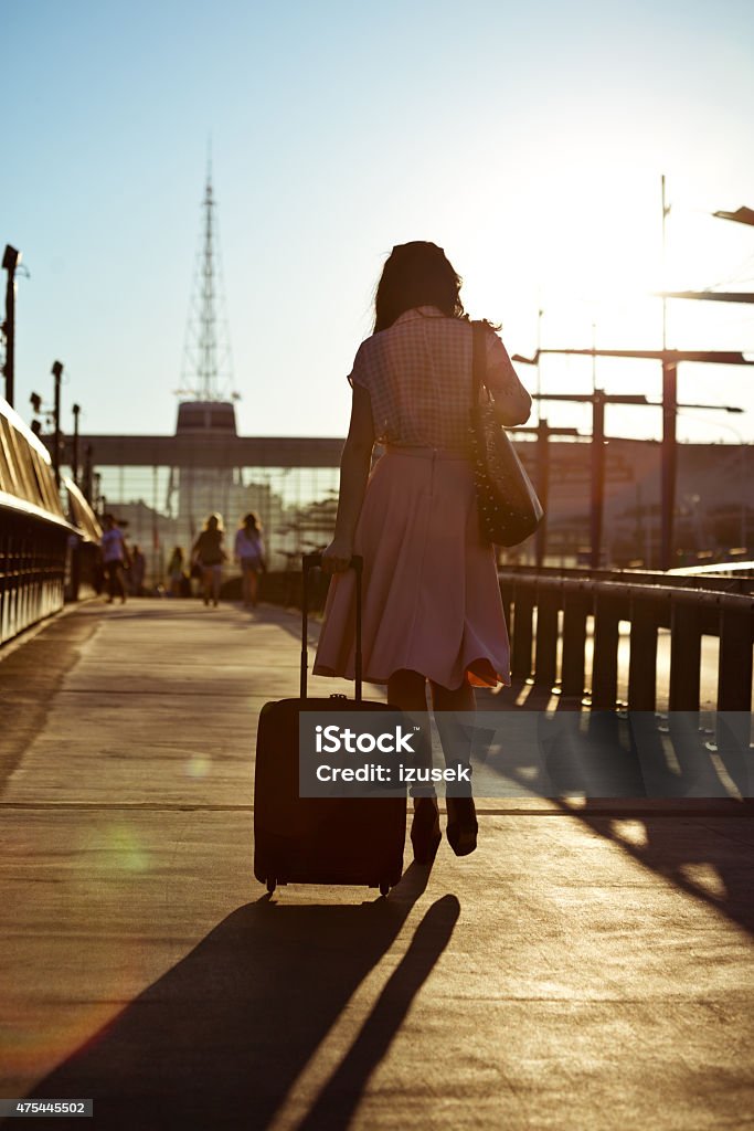 Young woman walking sidewalk and carrying suitcase Back view of young woman wearing blouse and skirt walking down the sidewalk in the city at sunset, carrying suitcase.  2015 Stock Photo
