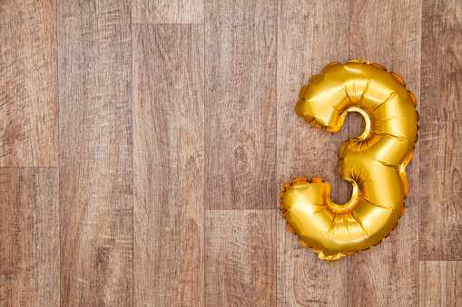 A gold foil number 3 balloon on a wooden background. The number is made from shiny golden foil and is inflated, it is on the right hand side of the image leaving copy space on the left of the image for your text or logo.