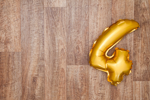 A gold foil number 4 balloon on a wooden background. The number is made from shiny golden foil and is inflated, it is on the right hand side of the image leaving copy space on the left of the image for your text or logo.