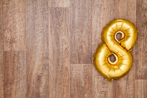 A gold foil number 8 balloon on a wooden background. The number is made from shiny golden foil and is inflated, it is on the right hand side of the image leaving copy space on the left of the image for your text or logo.