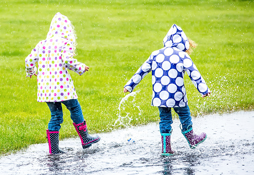 Rear view of two young girls (sisters) splashing through a large puddle in the rain. They are both wearing polka dot rain coats and rubber boots.