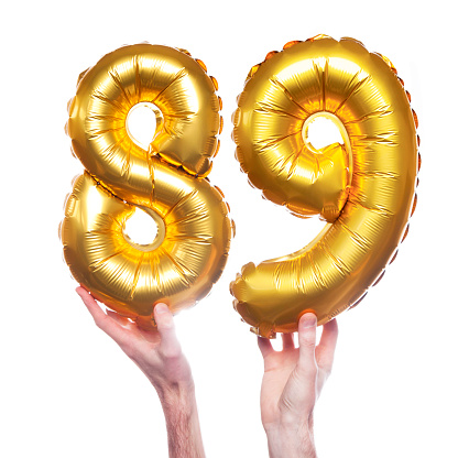 Gold foil number 89 balloons held by a caucasian male hand. The numbers are being held at the base. The numbers are made from shiny golden foil and is inflated. The background is plain white.