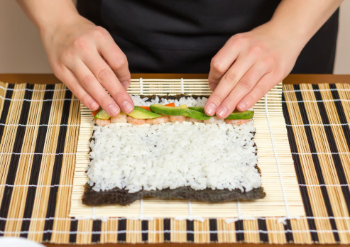 Hands of woman chef rolling up a japanese sushi with rice, avocado and shrimps on nori seaweed sheet