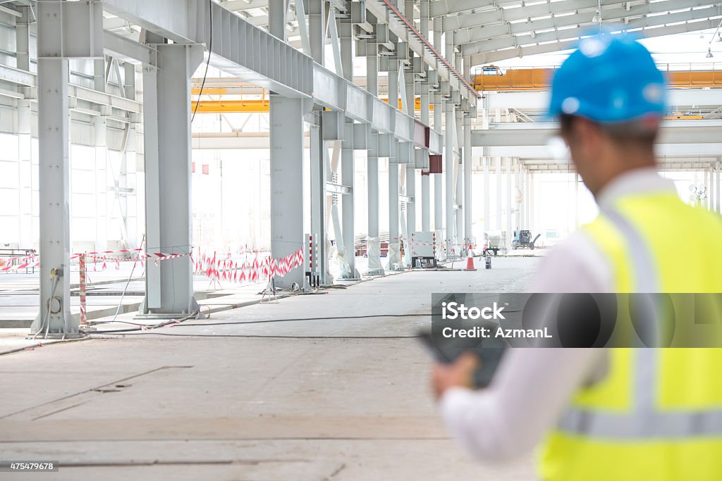 Construction Foreman Checking Progress Construction foreman on construction site, holding digital tablet. He is wearing reflective vest over a white shirt. We can see construction site in back, foreman is in front, defocused. He is looking away from the camera. Copy space. Construction Site Stock Photo