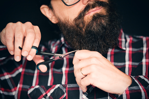A close up of a man holding scissors about to cut his long hipster beard off.