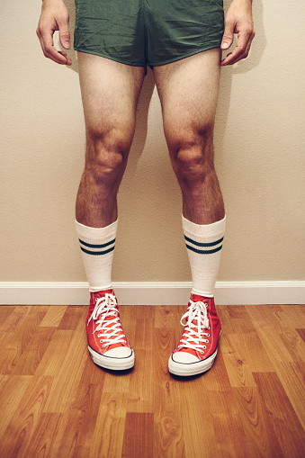 A man in 1970's - 1980's styled gym and sports active wear clothing wears high top canvas sneakers, short shorts, and knee high striped socks, sporting a glaring white ugly farmers tan line on his legs.  Vertical image, focus on the tennis shoes.